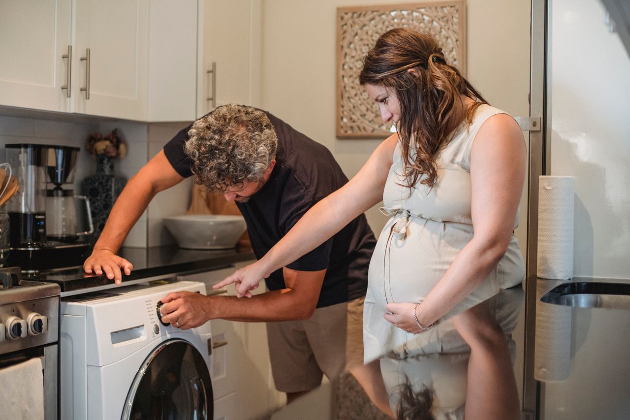 Bearded man with gray curly hair assisting pregnant wife to turn on washing machine