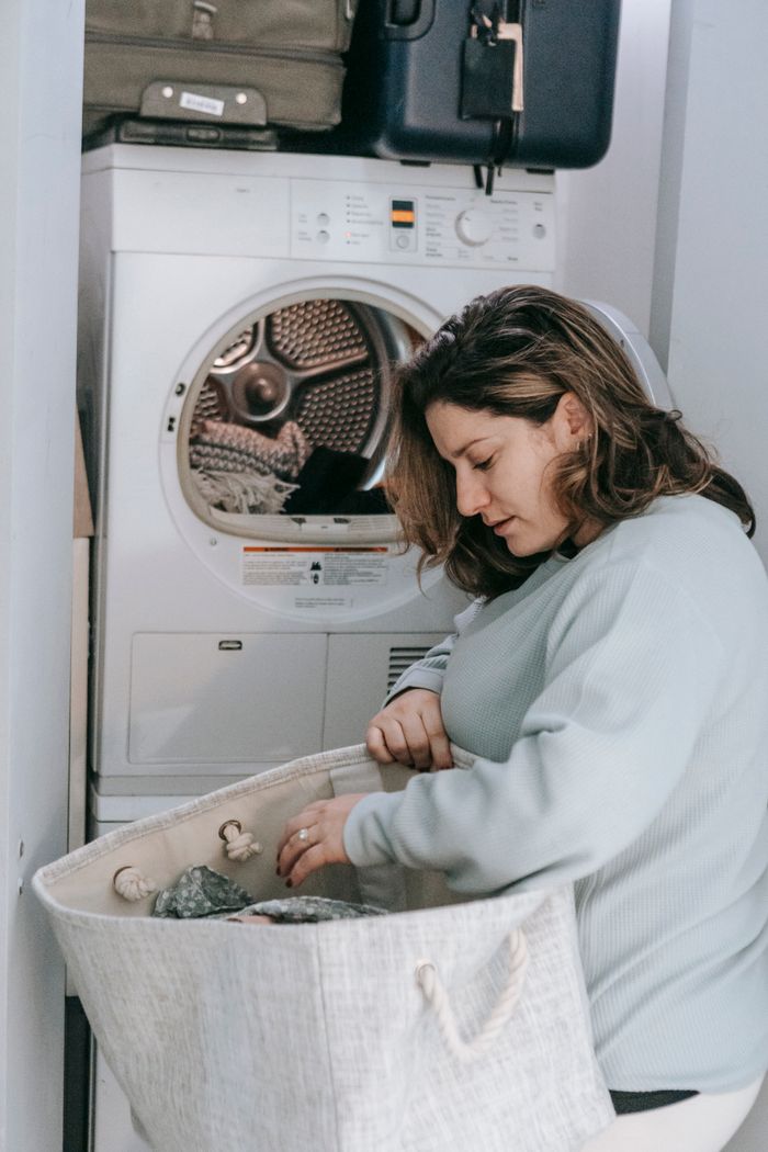 services-07 Side view of serious female with bag of dirty clothes loading modern washing machine while sitting in light washroom at home